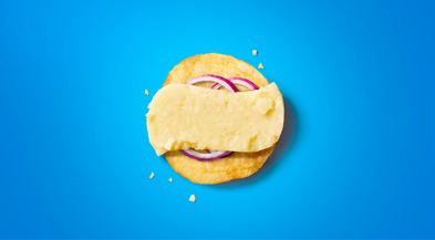 A single tortilla chip topped with a slice of cheese and red onion, on a vibrant blue background.