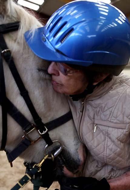 A person in a blue helmet gently nuzzling a white horse's face, showing an affectionate interaction.
