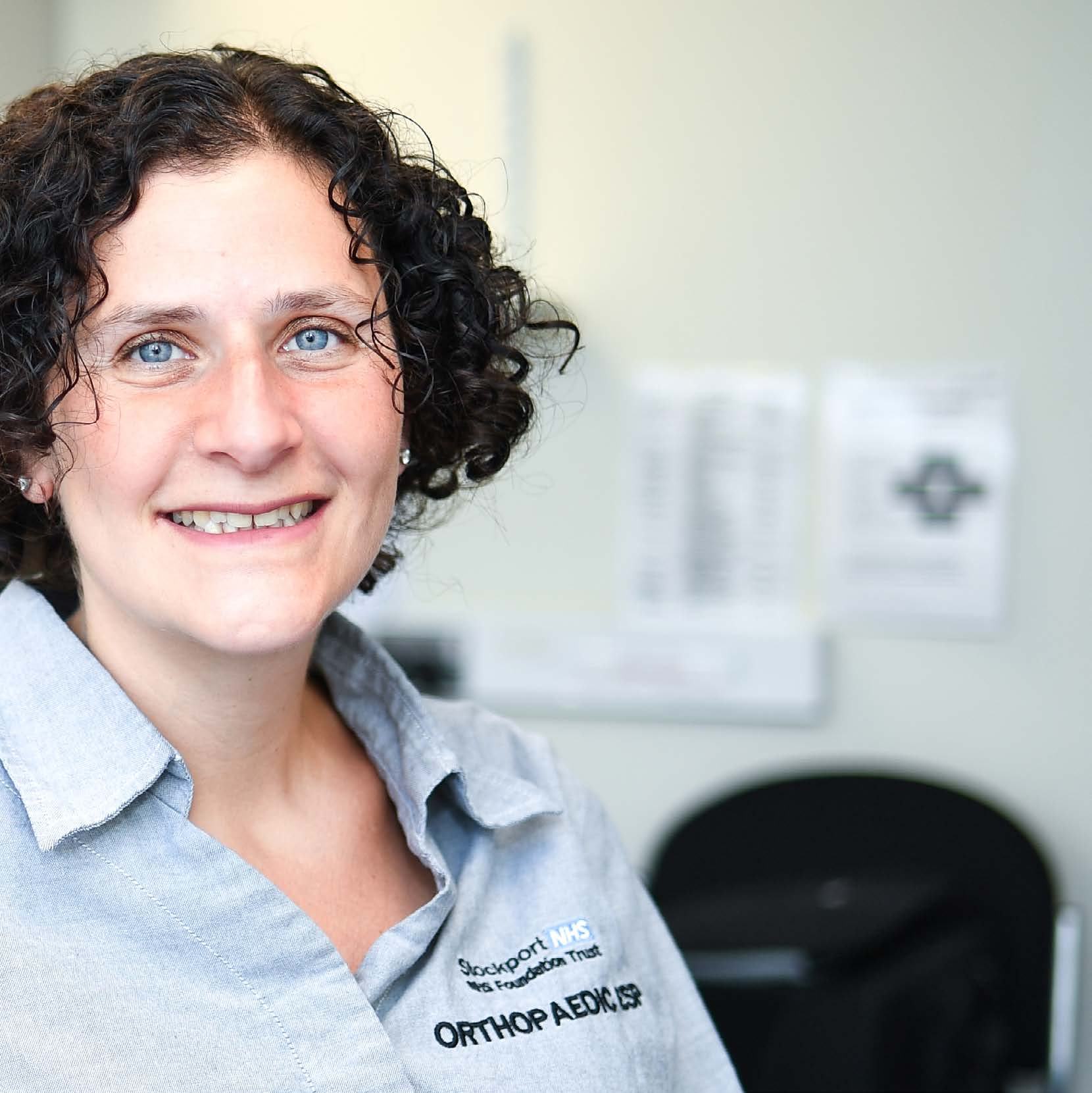 Headshot of a smiling woman with curly hair, wearing a shirt that says "Stockport NHS Foundation Trust ORTHOPAEDIC, ISP".