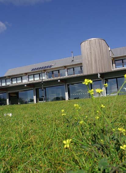 Modern building with glass facade, wooden circular section, and solar panels on the roof, viewed from a grassy field.