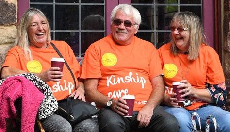 Three people in orange "Kinship Care" t-shirts laughing and holding coffee cups on a bench.