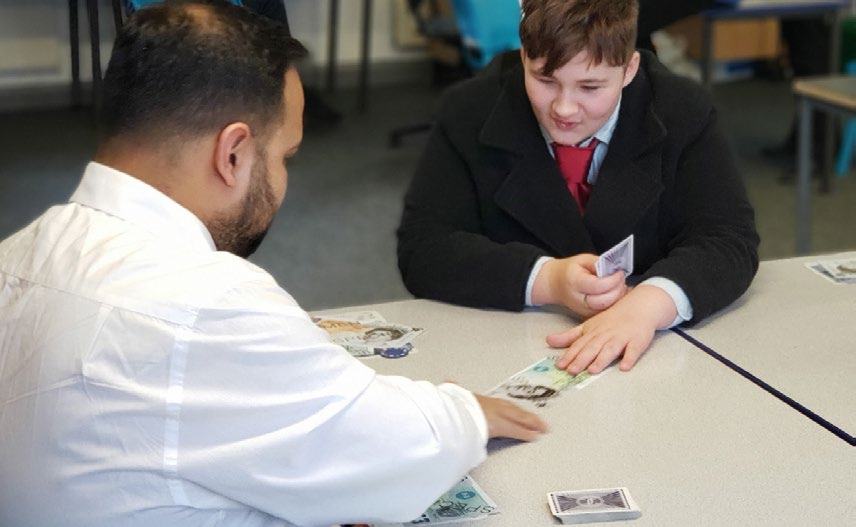 An adult and a young person sitting at a table, engaged in a game or lesson involving paper money (pounds) and playing cards.