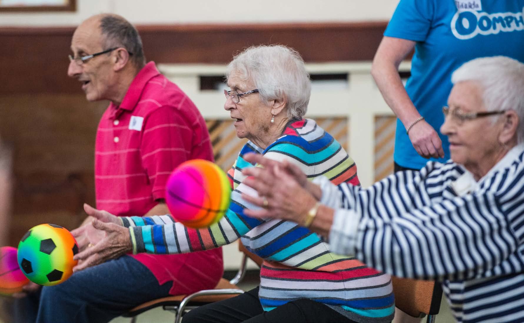 Elderly individuals engage in a seated exercise class, holding and passing colorful balls. A person in the background wears a shirt with the 'Oomph!' logo.