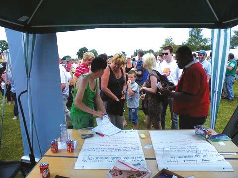 People gathered under an outdoor canopy, interacting around a table with large sheets of paper displaying handwritten text and questions, likely at a community event.