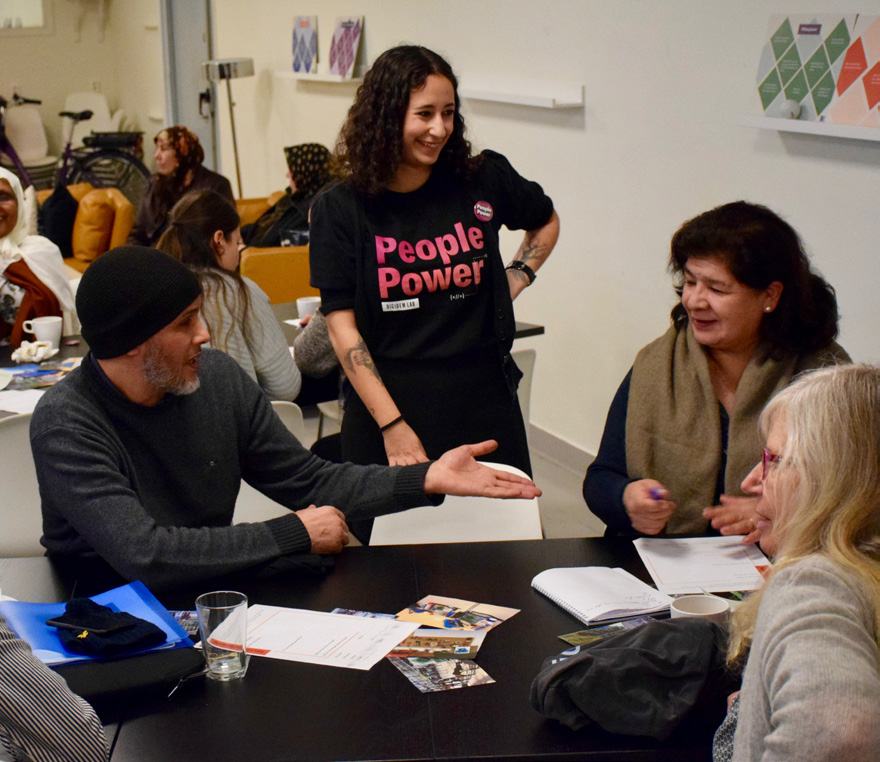 A group of people in a lively discussion at a meeting table. One woman stands, wearing a "People Power" shirt.