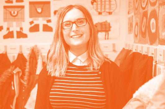 Smiling woman in glasses and striped shirt stands in a classroom with children's artwork.