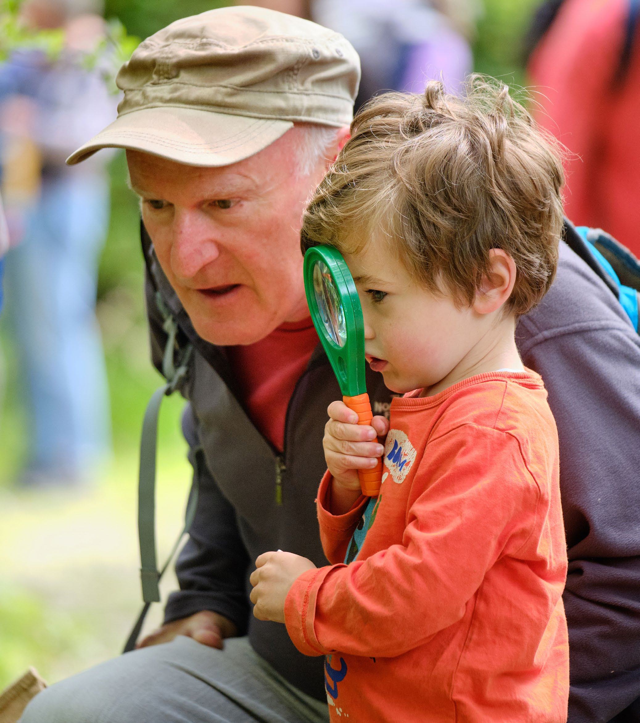 An adult man and a young child outdoors, with the child holding a magnifying glass to their eye.