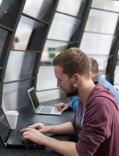 Man with a beard typing on a laptop in a modern workspace, with another person working in the background.