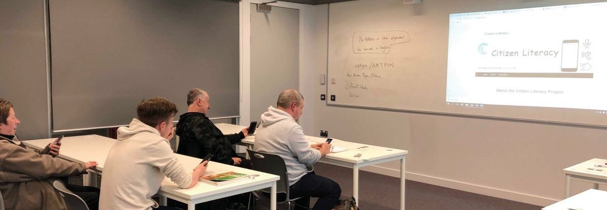 Classroom of adults using smartphones, with "Citizen Literacy" project displayed on a projector screen.