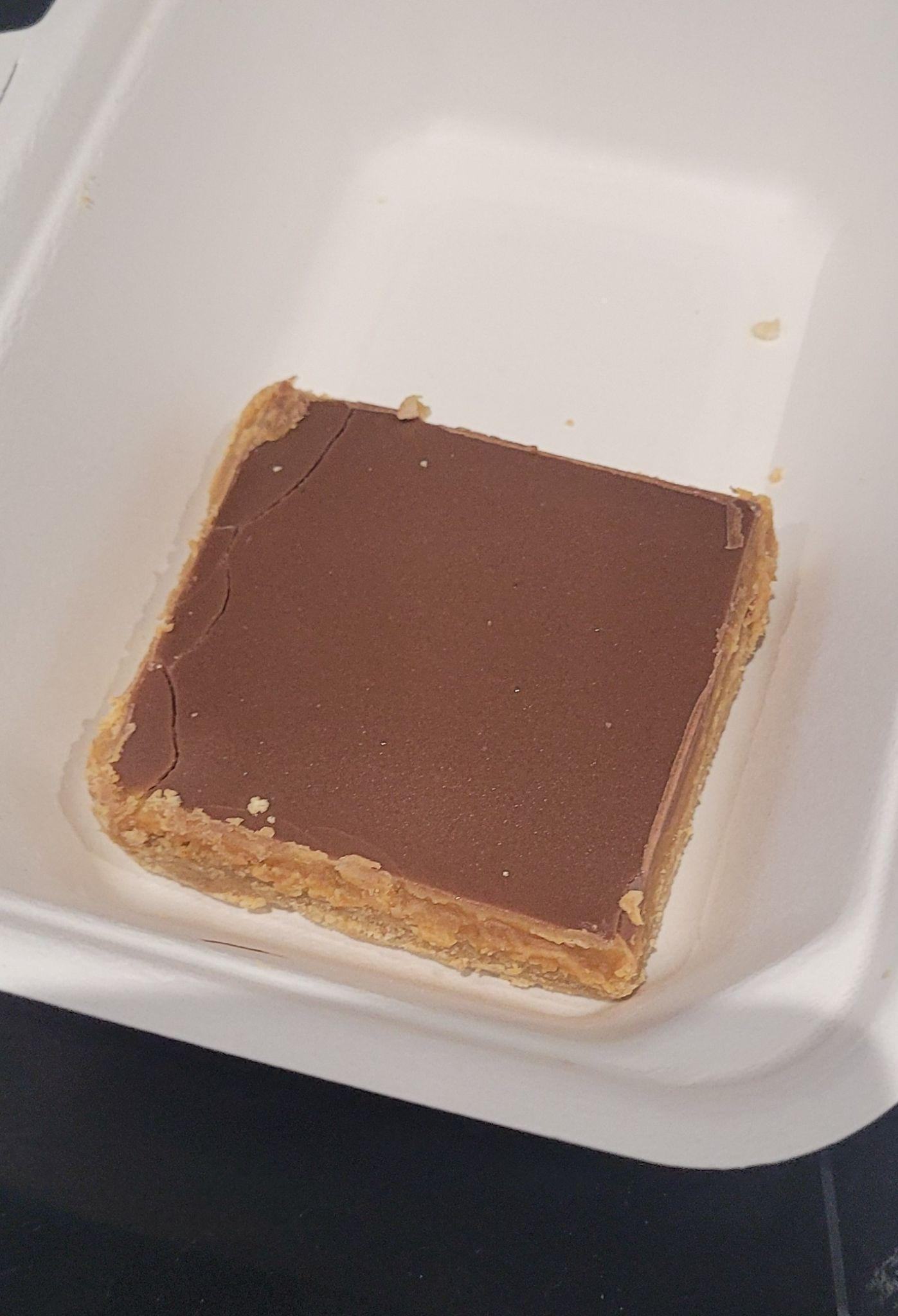 A square chocolate-topped biscuit or dessert bar in a white, disposable container.