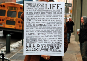 A person holding a large poster with a detailed motivational quote about living life, work, and passion, on a city street.