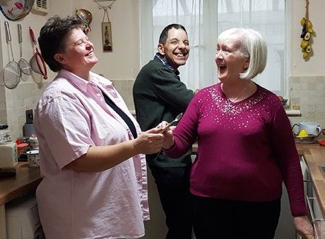 Three people in a kitchen laughing together, including a woman in a pink shirt and an older woman.