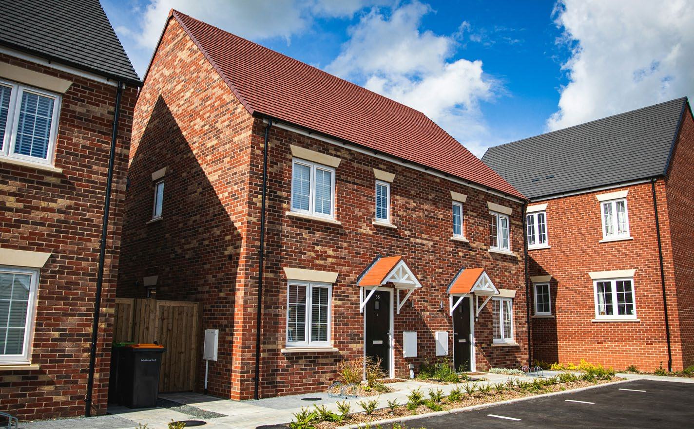 Row of newly built brick houses with red and grey tiled roofs, white windows, and small gardens under a blue sky.