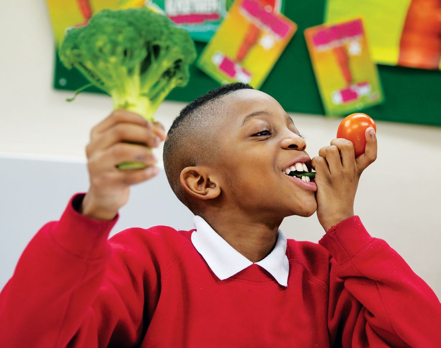 A smiling boy in a red sweater holds a broccoli floret in one hand and a tomato in the other, illustrating children's engagement with healthy eating.