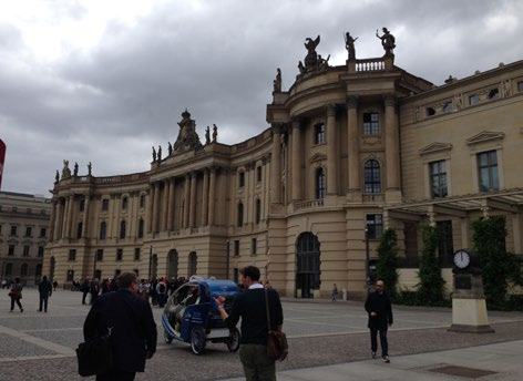 A grand, classical-style building with ornate sculptures on the roof and columns, overlooking a large paved plaza with people and a small vehicle.