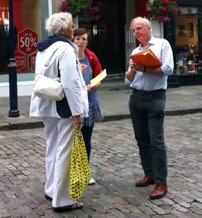 Three people, two older and one younger, conversing on a cobblestone street; one man holds a folder and takes notes.