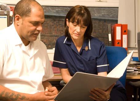 A nurse in uniform and a man in a white shirt sit at a desk, both looking at a document held by the nurse, suggesting a consultation.