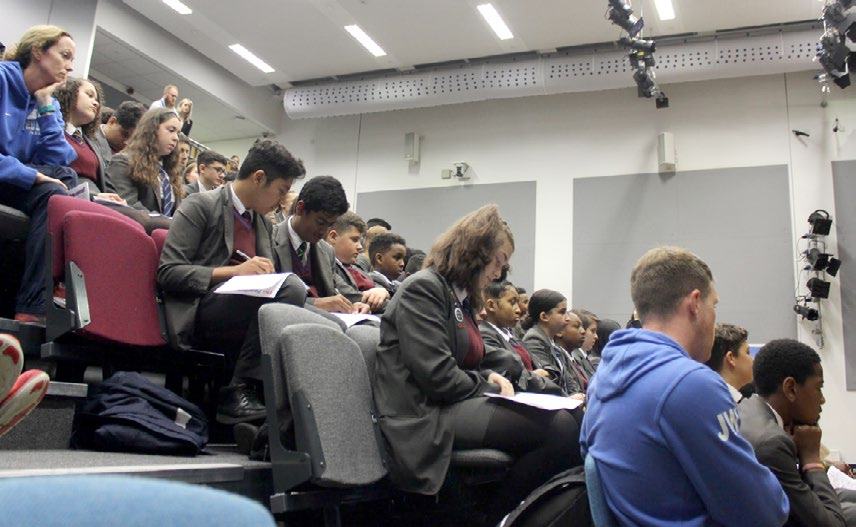 Students and a few adults seated in a lecture hall, with many students in uniform taking notes or listening to a presentation.