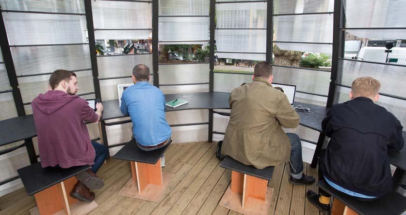 Four men working on laptops at individual desks inside a semi-circular, translucent outdoor workspace.