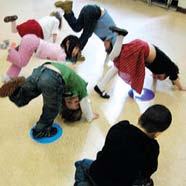 Several young children in colorful clothing performing a playful bending exercise or yoga pose on the floor, some using small colored mats.