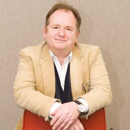 Headshot of a smiling man in a beige jacket and white shirt, leaning on a red chair back.