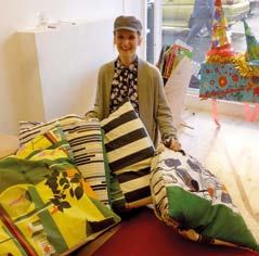 A woman in a cap smiling behind a large pile of colorful, patterned cushions, with a festive piñata-like decoration visible in the background.