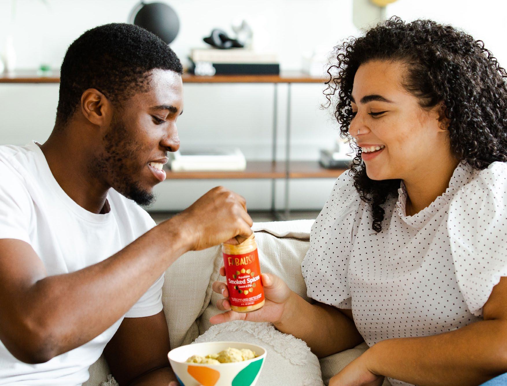A man opening a jar of F. Radish Smoked Bacon snacks while sitting on a couch with a woman.