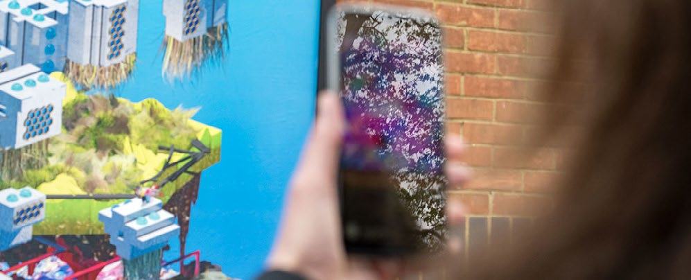 A person holding a smartphone takes a picture of a colorful mural depicting floating structures and landscapes.