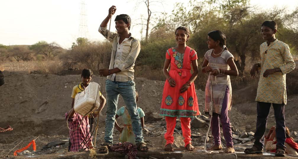 Group of adults and children in a rural setting, with a man operating a simple well mechanism and others observing.