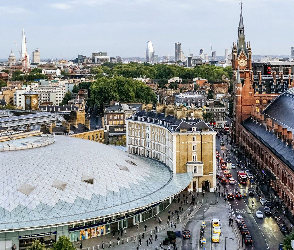 Aerial view of a city, showing a modern building with a unique roof, St Pancras station, and other urban structures with traffic below.