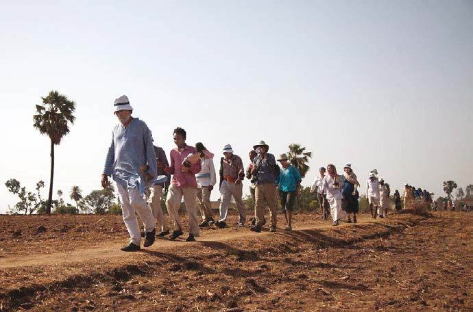 A group of people walking along a dusty path in a rural landscape under a clear sky with scattered palm trees.