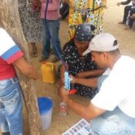 A man demonstrates a portable water filter to others on the ground, with a yellow jerry can nearby.