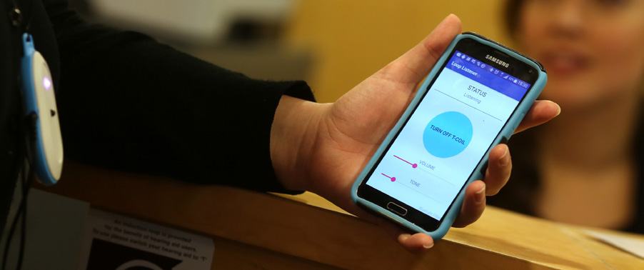 A hand holding a smartphone displaying the "Loop Listener" app interface, showing "STATUS Listening" and controls for volume, tone, and T-coil activation.