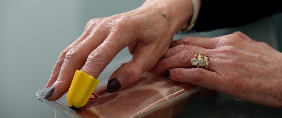 Close-up of hands using a yellow gripper tool to seal a plastic bag containing sliced meat, demonstrating an assistive device.