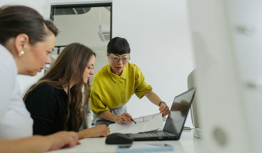Three women reviewing documents and a laptop, with one pointing at a technical drawing or blueprint.