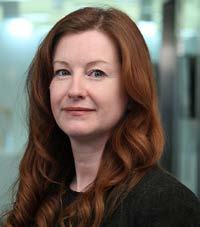 Headshot of a woman with long red hair, looking slightly right with a gentle smile, in a blurred office background.