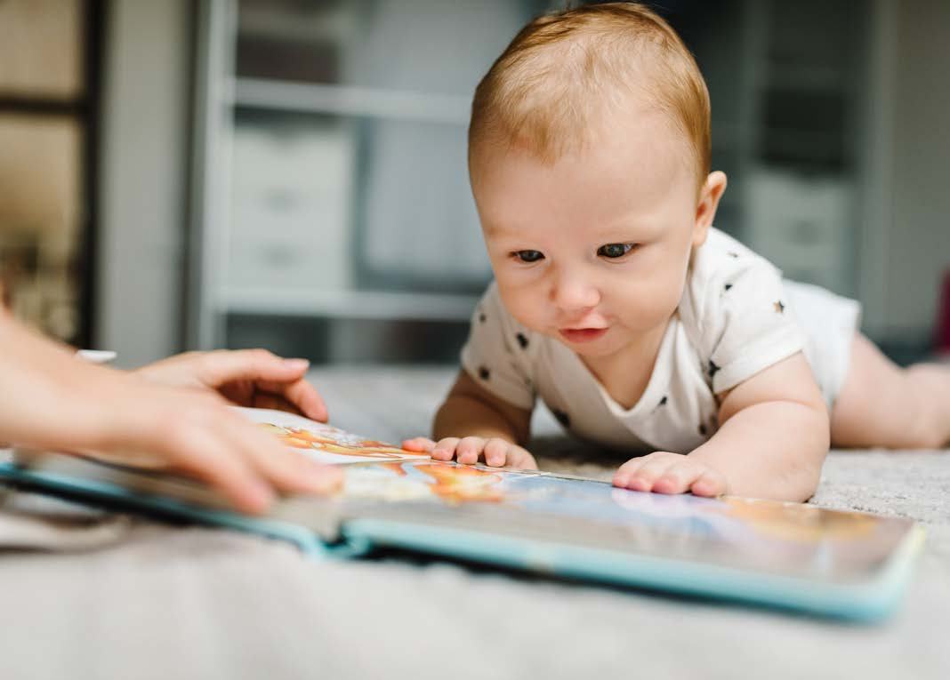 A baby lies on a rug, looking intently at an open picture book, with an adult's hands visible.