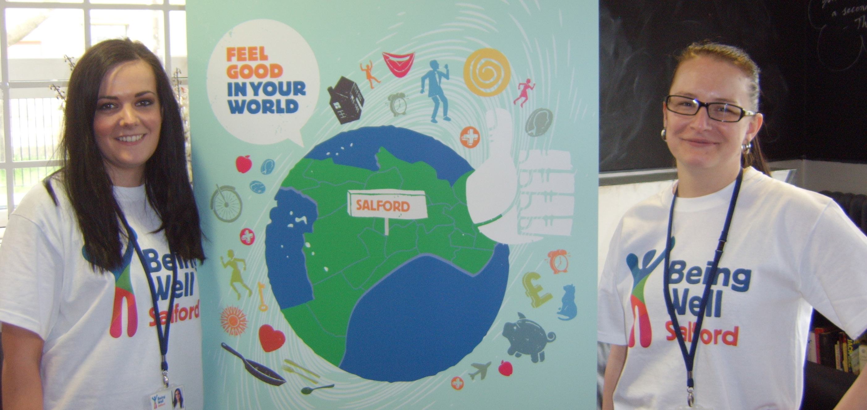 Two women in 'Being Well Salford' t-shirts stand in front of a poster with a map of Salford and icons representing well-being activities.