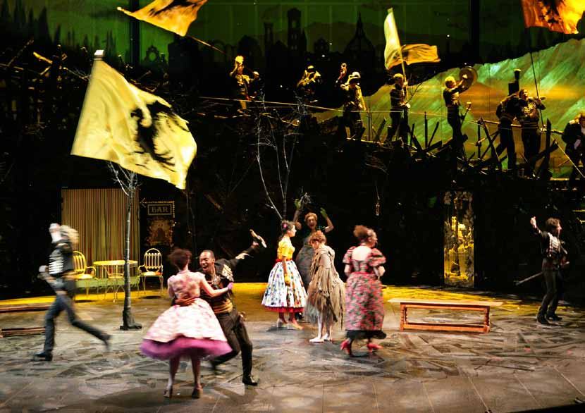 A theatrical stage scene with multiple actors in period costumes, some dancing, some holding flags, against a dark, dramatic backdrop.