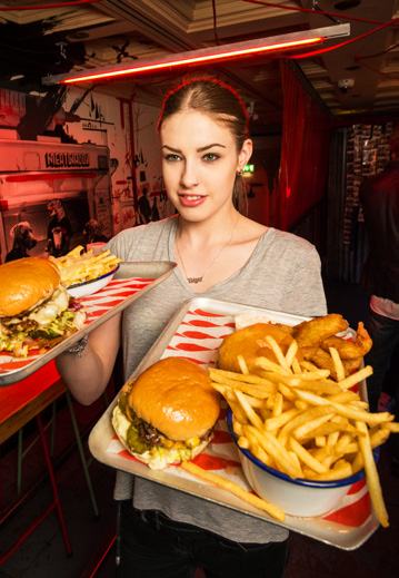 Female server holding two trays of food, including burgers, french fries, and fried chicken, in a dimly lit restaurant.