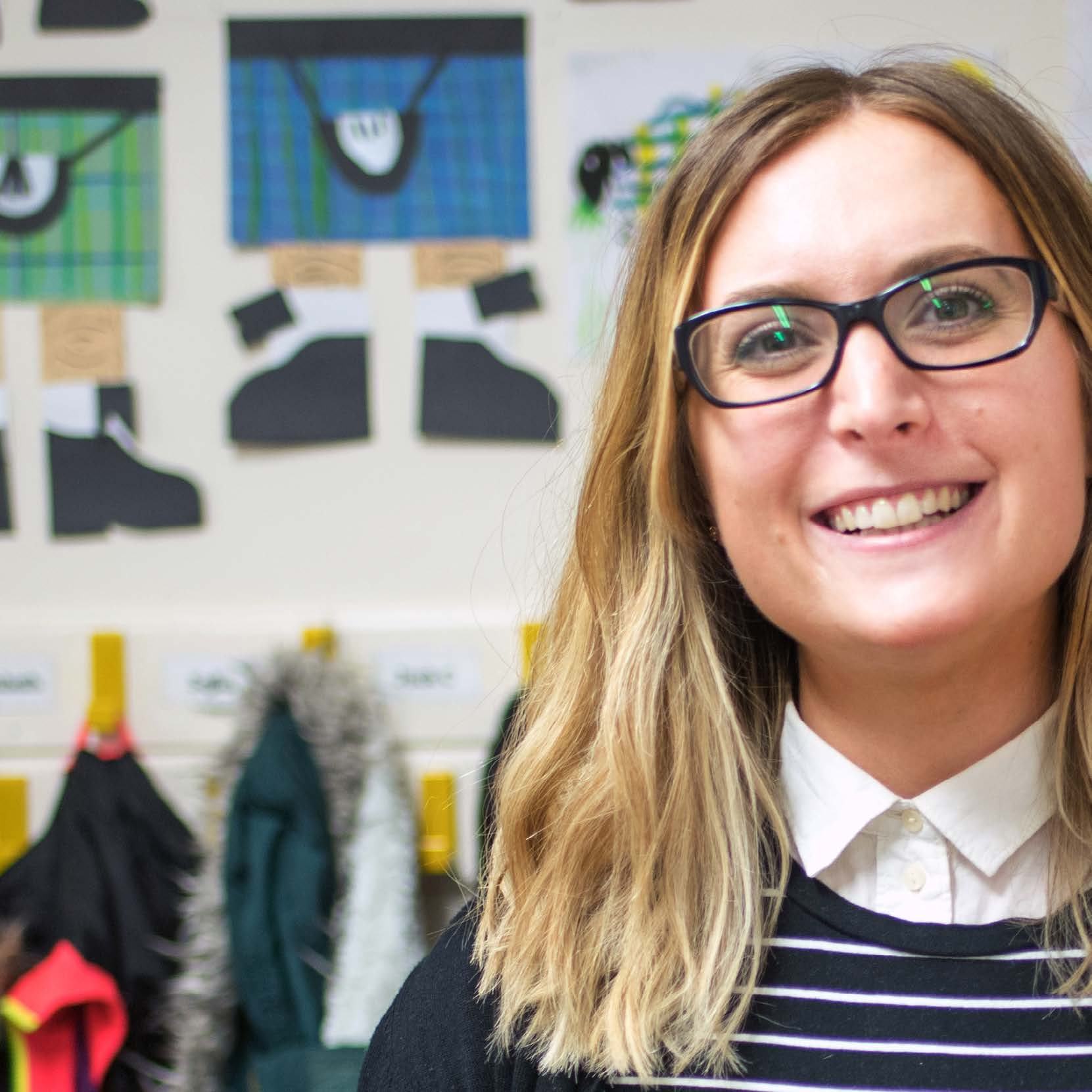 Smiling woman with glasses in a classroom setting, with colorful children's artwork displayed on the wall behind her.