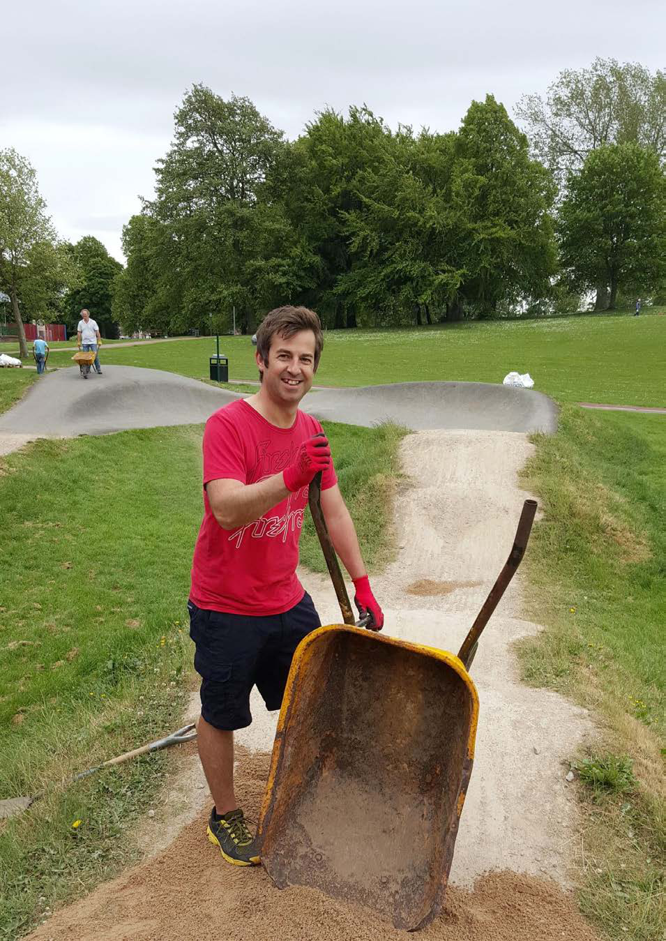 Man in red shirt and gloves smiling next to a yellow wheelbarrow full of sand on a dirt track in a park, with another person in the background.