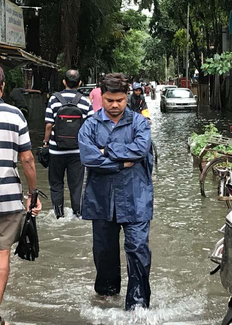 Man in a blue raincoat walking through a flooded street, with other people and submerged vehicles in the background.