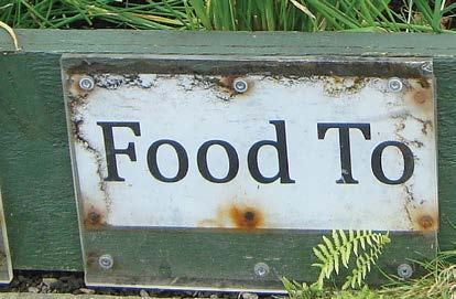Weathered white sign with "Food To" written on it in black letters, partially obscured, in a garden setting.