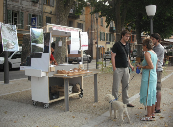 People observing an outdoor stand with displays and a physical model, possibly an educational or community engagement booth.