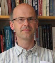 Headshot of a man with glasses and a striped shirt in front of a bookshelf.