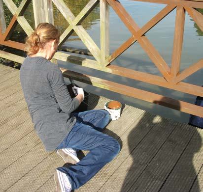 Person in jeans and a gray shirt sitting on a wooden deck, painting the railing of a bridge over water.