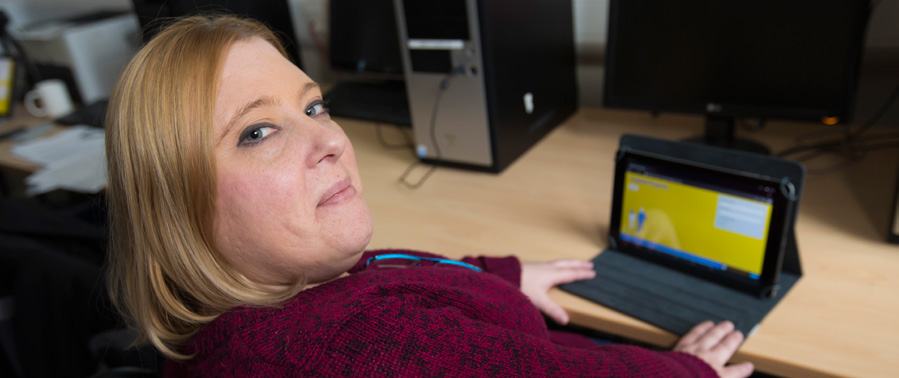 A woman with blonde hair in a red sweater looks at the camera while using a tablet at a desk, with a desktop computer behind her.