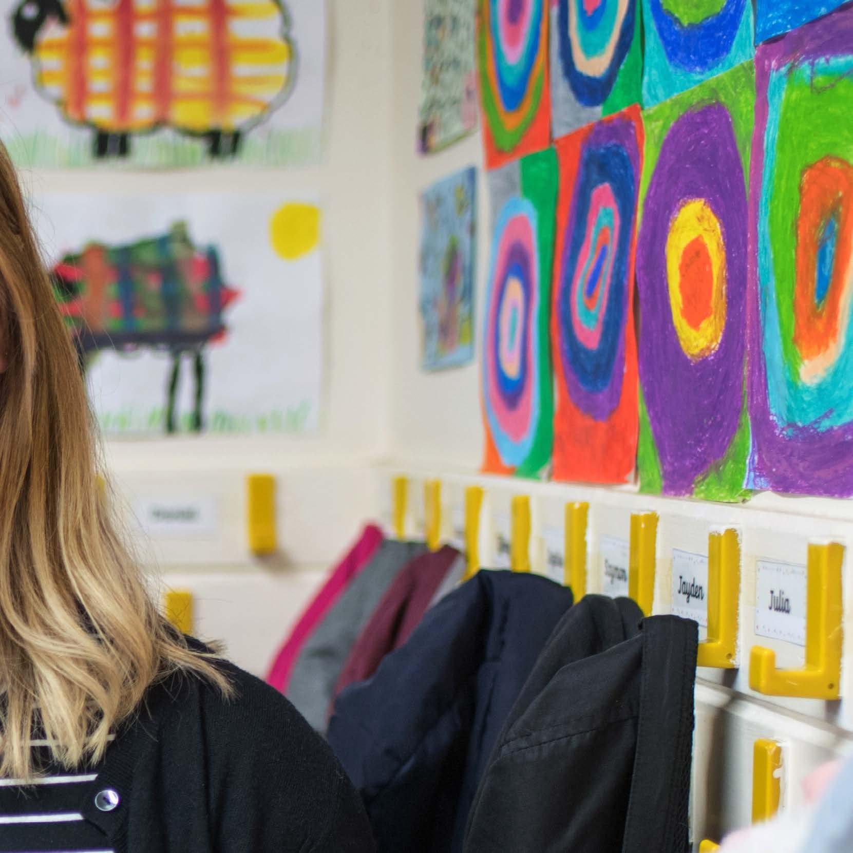 Classroom wall with colorful children's artwork and coats hanging on yellow hooks, with student names visible.