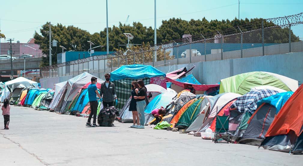 Long row of diverse tents set up in an outdoor camp, with people standing among them, suggesting a temporary settlement or shelter.
